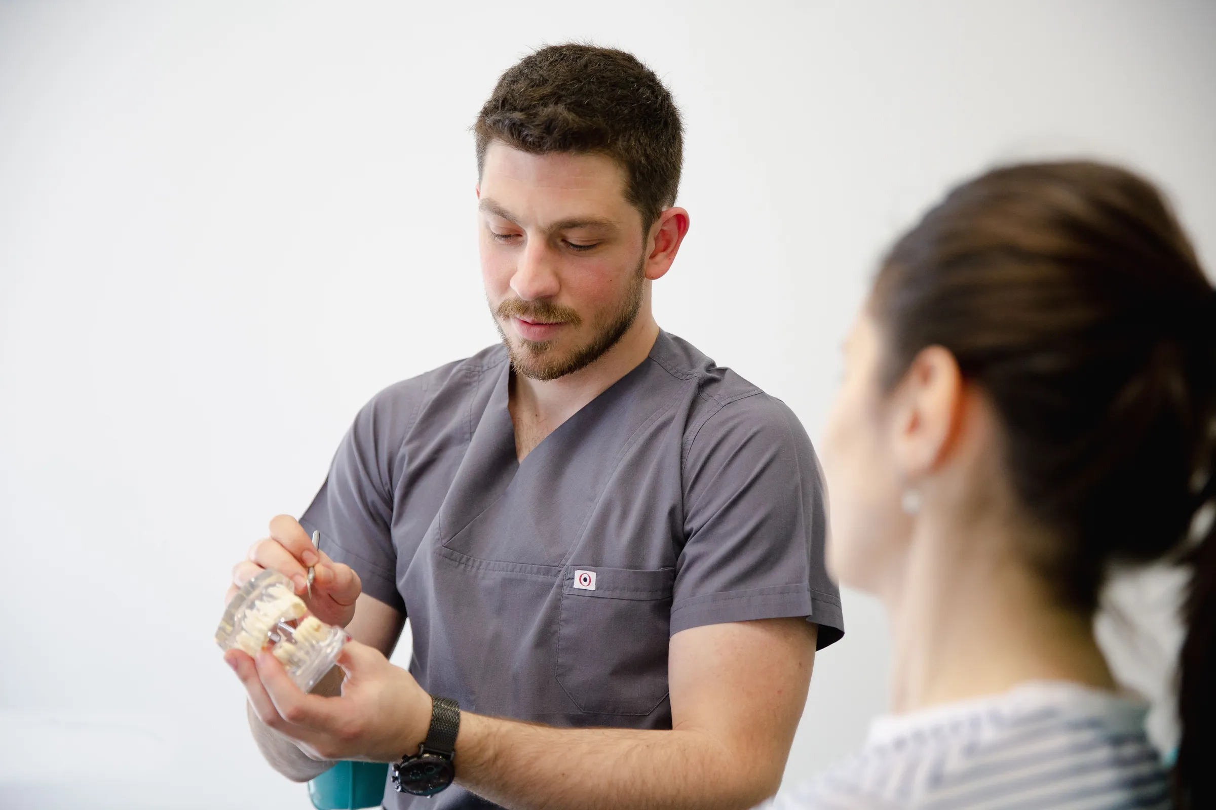 A clinician in gray scrubs holds a model of teeth while explaining treatment to a patient